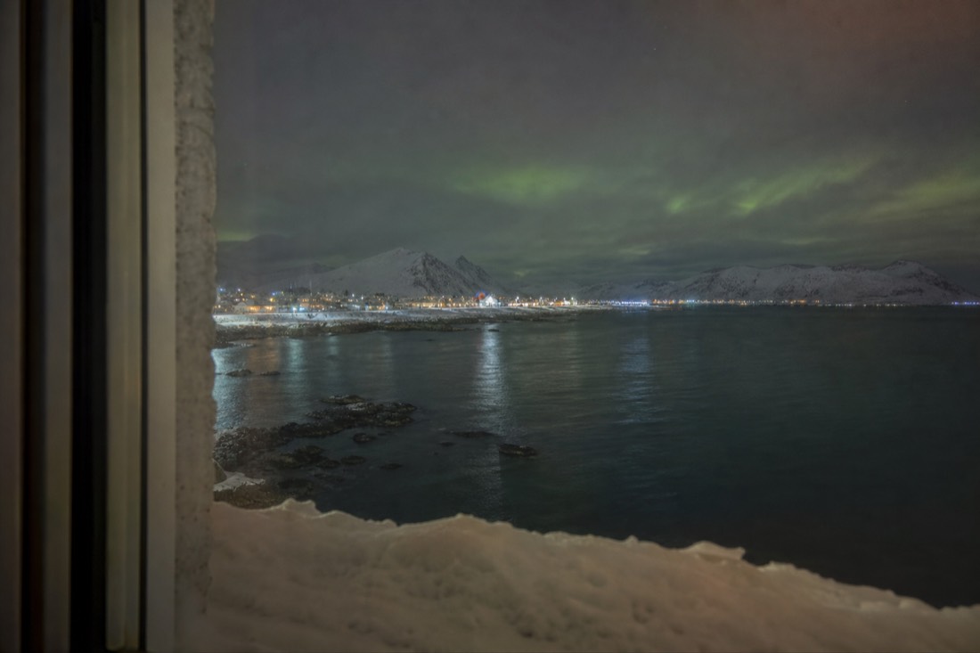 Hotel window at night in an Arctic town with snow on the sill and faint green light in the sky