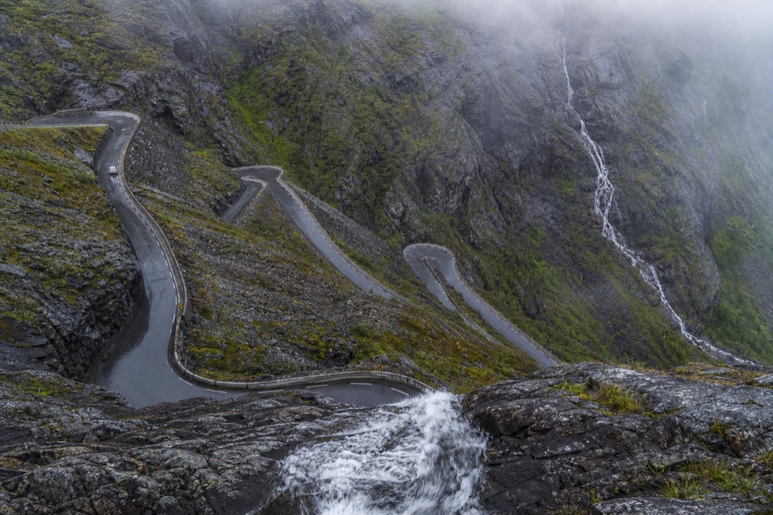 Narrow Norwegian mountain road with hairpin bends cut into rock, guardrails and a waterfall on the far slope