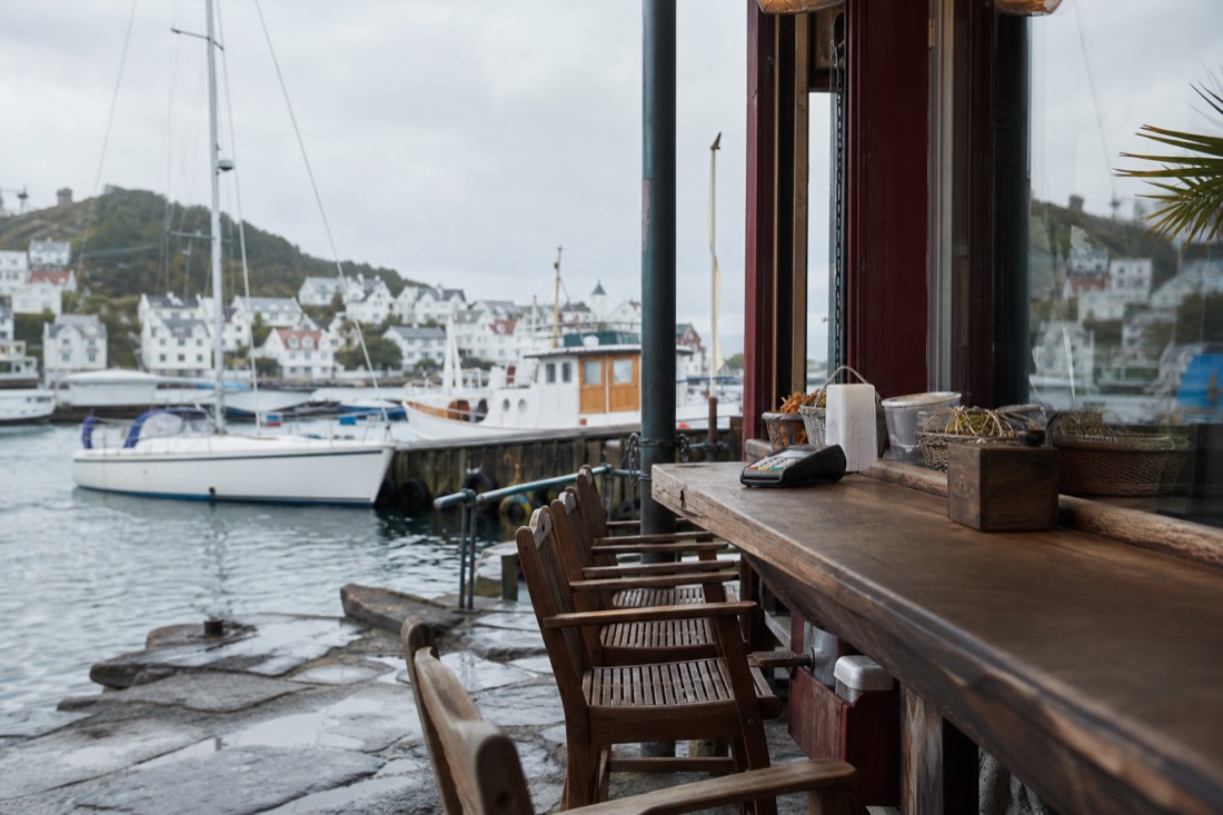 Waterfront cafe terrace in a Norwegian town with wooden chairs, a card reader on the counter and boats moored behind