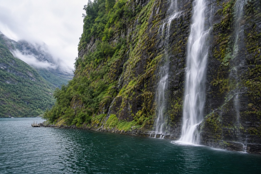 Waterfall spilling down a mossy cliff directly into deep green fjord water beside a small jetty