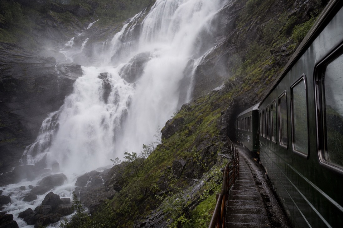 Green mountain railway carriage stopped beside a thundering waterfall with passengers on the platform in spray