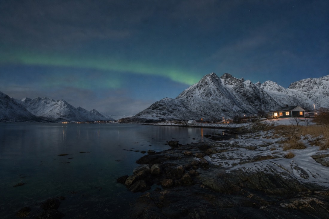Faint green aurora band above a snow-covered fjord shoreline with dark peaks and a lit cabin window