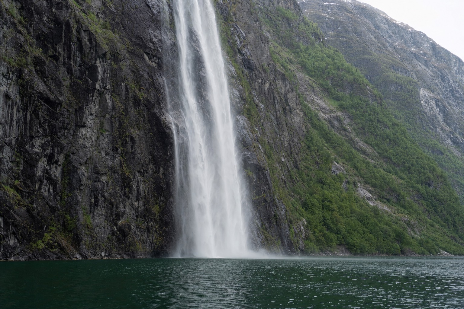 A narrow Norwegian fjord with a tall waterfall dropping directly into deep green water between sheer cliff walls