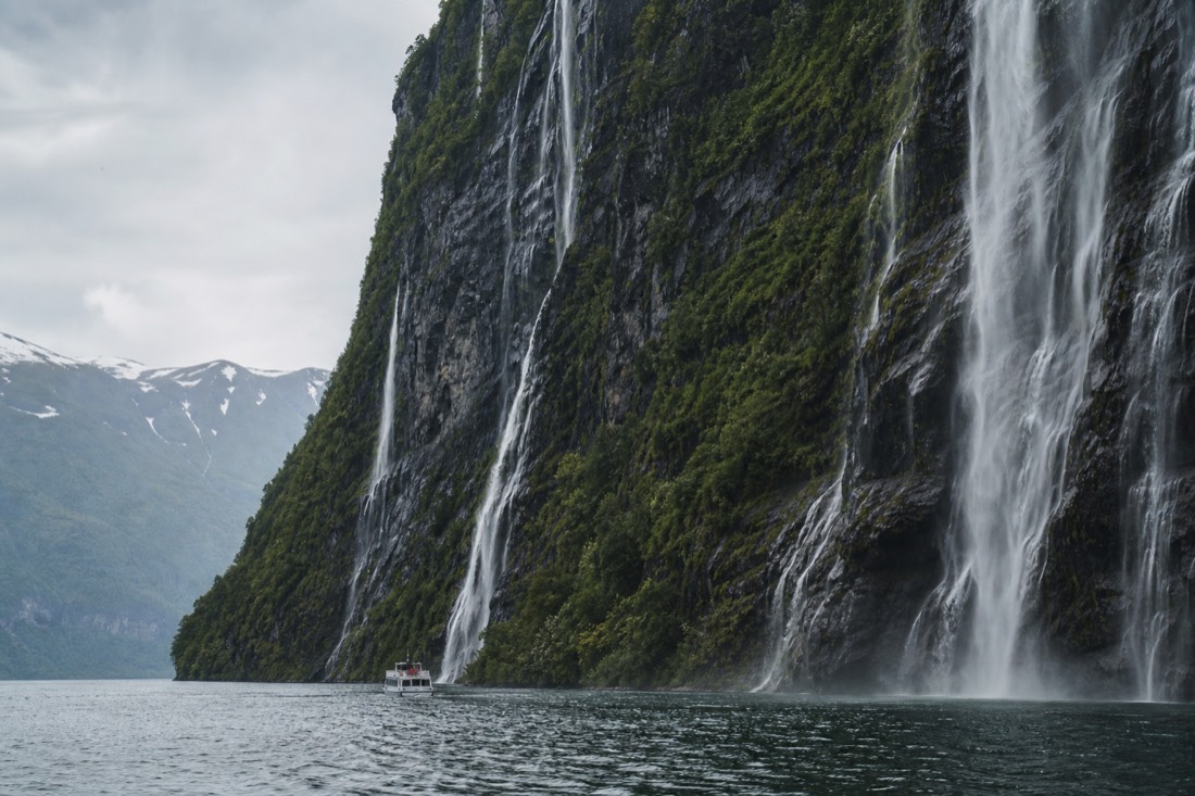 A sightseeing boat dwarfed by sheer green fjord walls with multiple waterfalls falling into the water beside it