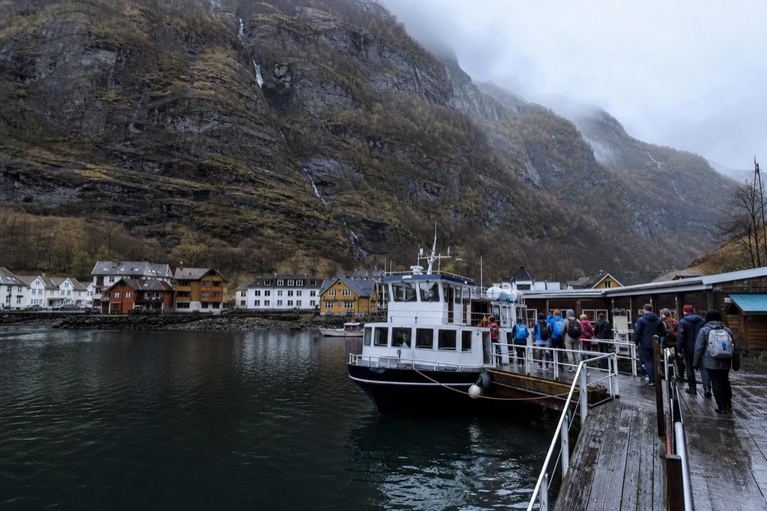 Passengers boarding a small sightseeing vessel at a wooden fjord quay with steep cliffs rising behind the village