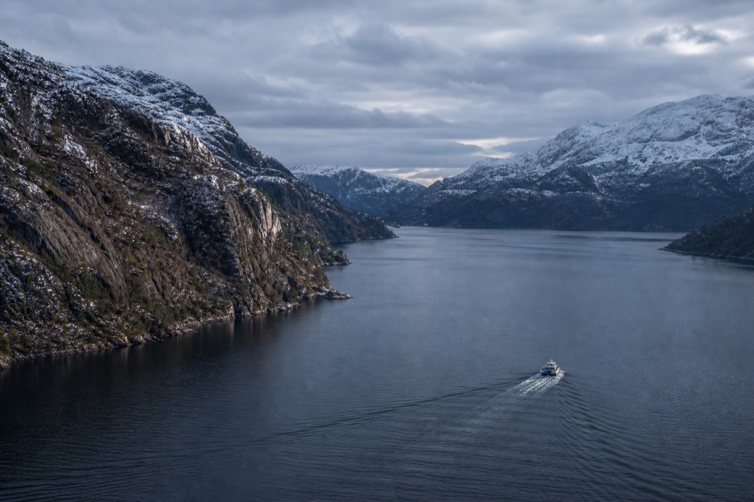 A winter fjord near Bergen with snow on the upper cliffs, dark water and a small vessel leaving a long wake