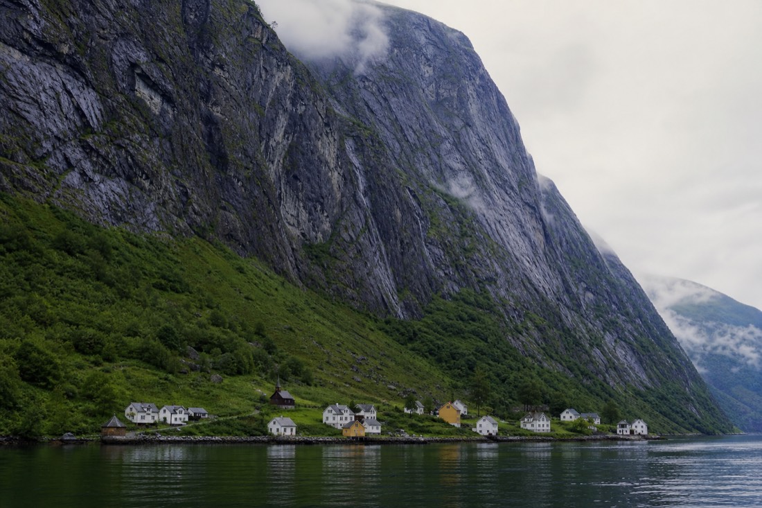 A cluster of white and ochre houses with a tiny wooden church on a narrow green shelf beneath towering fjord cliffs