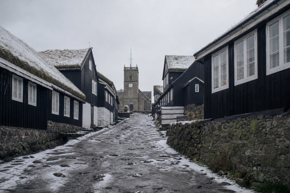 A wide unpaved street of dark weathered timber houses under snow with a stone church tower at the end