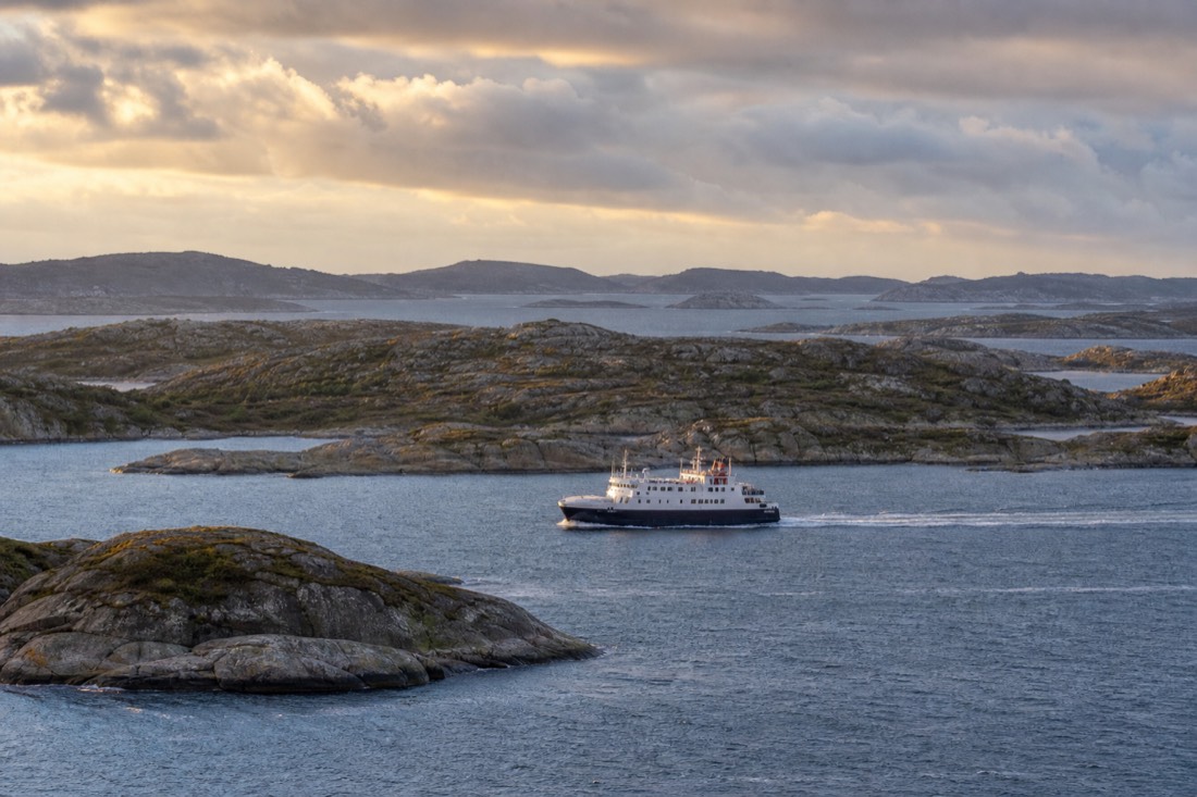 A coastal ferry cutting between small rocky islands on the Norwegian west coast in low golden evening light
