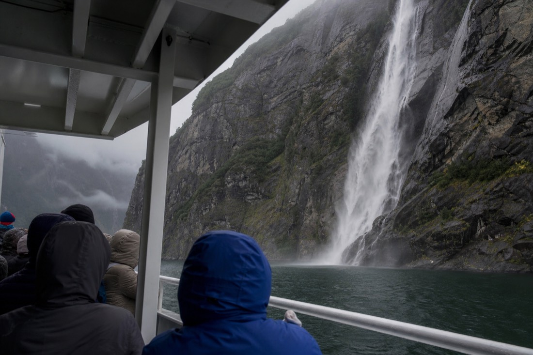 Passengers on the covered deck of a small fjord cruise boat watching a waterfall pour down a wet cliff face