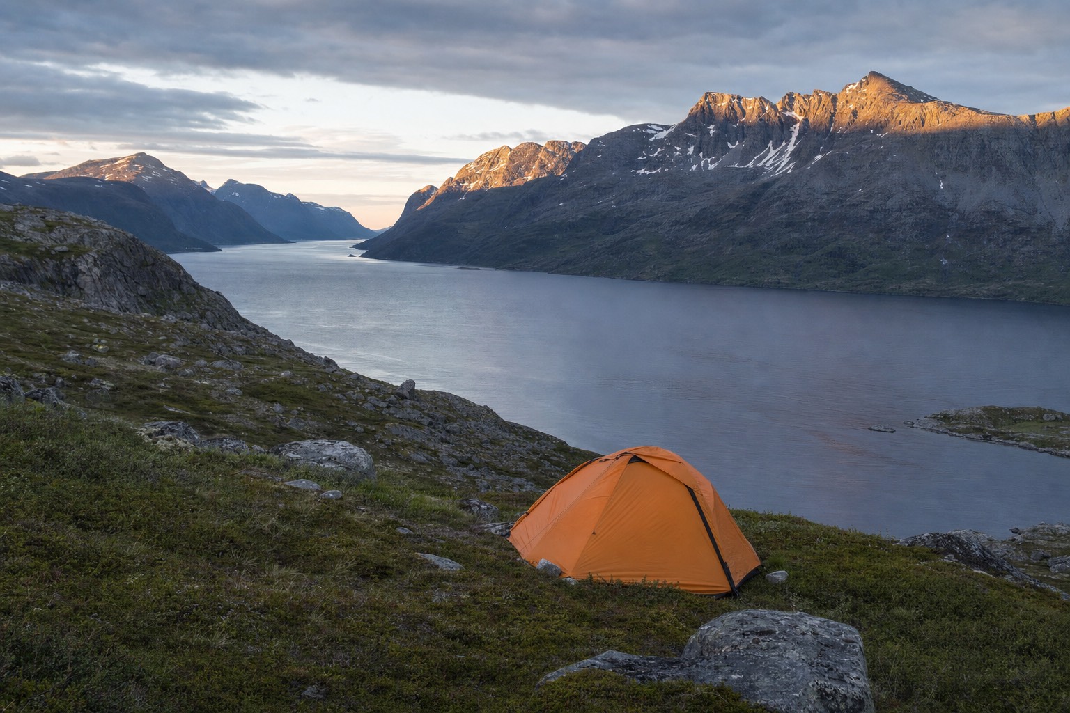 Small orange tent pitched on open moorland above a Norwegian fjord with mountains catching late evening light