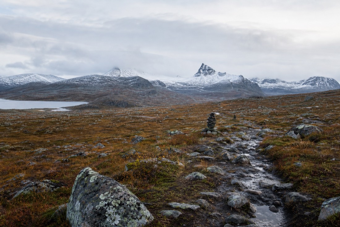 Worn hiking path crossing open Norwegian heath with cairns marking the route toward distant snow-capped peaks