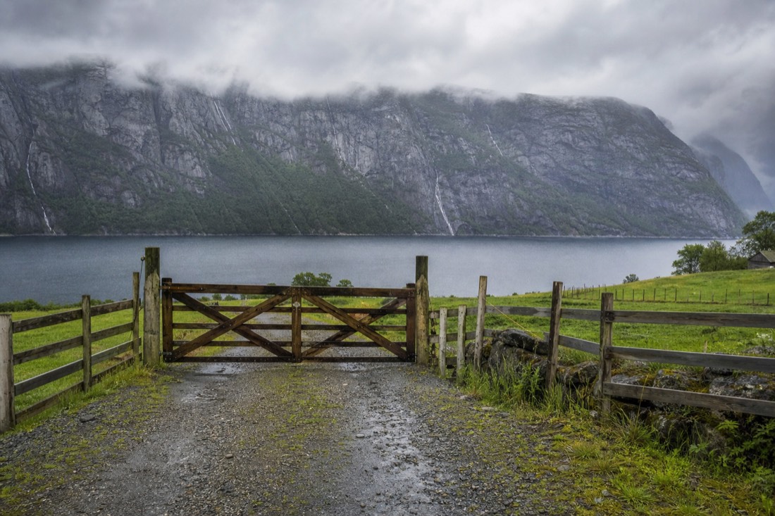 Wooden farm gate on a gravel track between fenced meadows with a fjord and mountain wall beyond