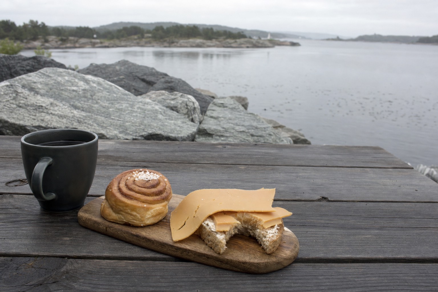 Open Norwegian sandwich with brown cheese and a cinnamon bun on a wooden table beside coffee