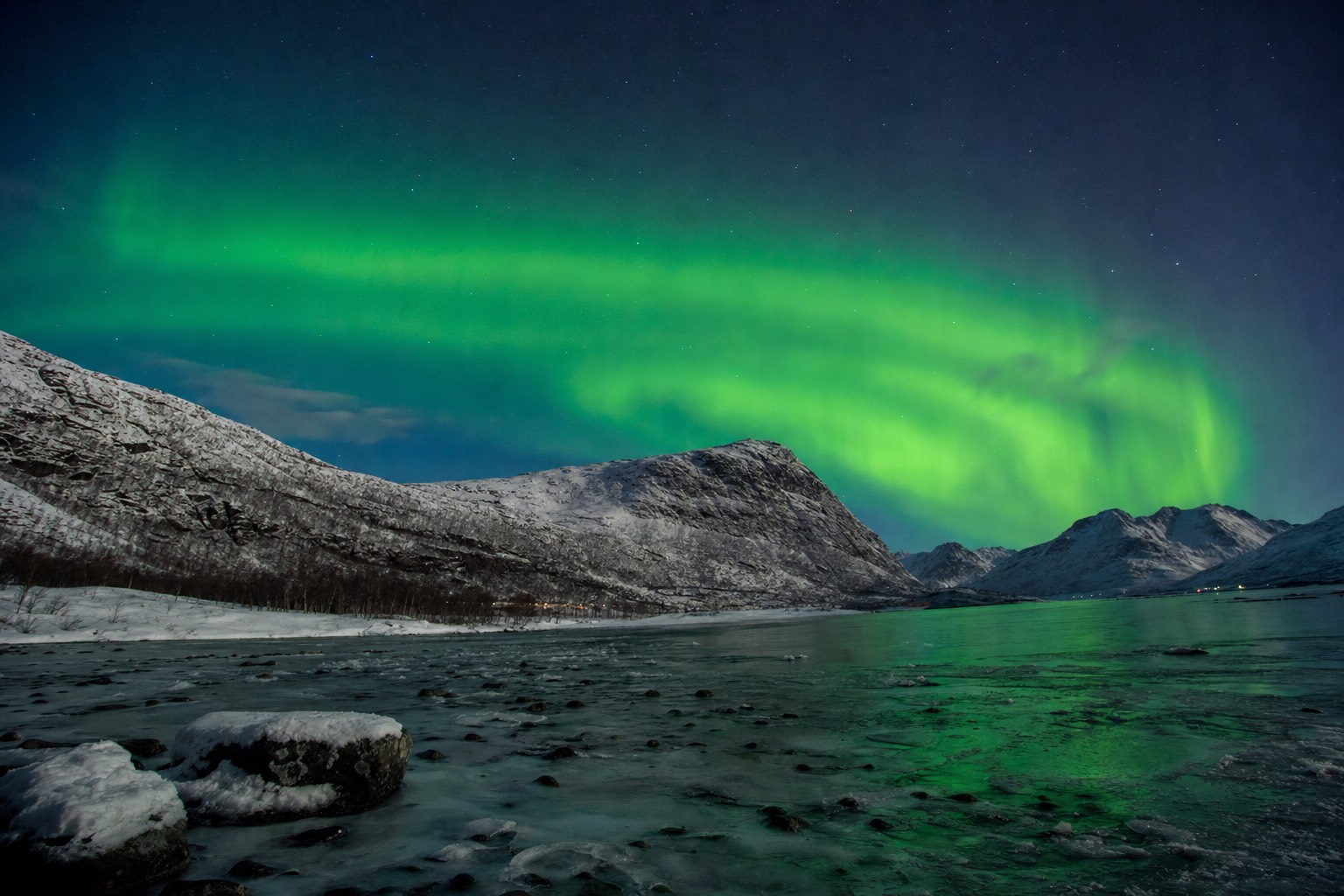Green aurora arc over a snow-covered Arctic valley with a frozen lake in the foreground