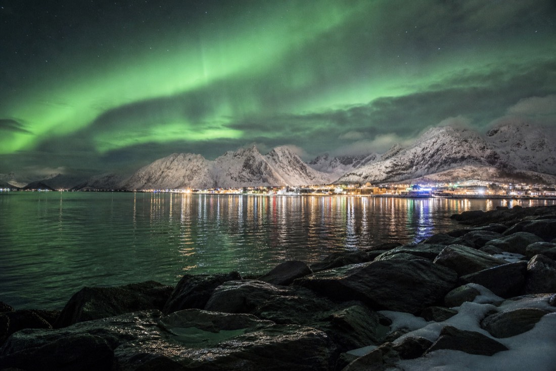 Small Arctic town lit at night beneath a green aurora with snowy mountains ringing the harbour