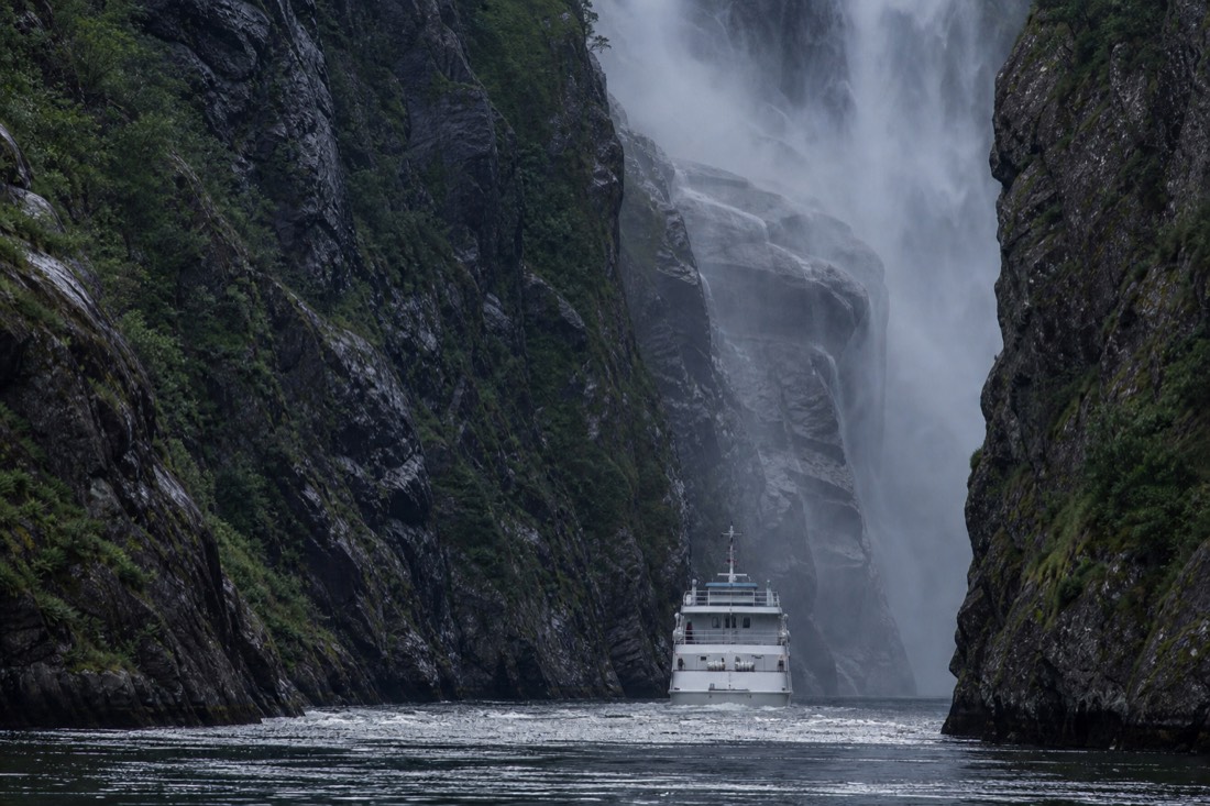 A white fjord cruise boat nosing toward a waterfall in the narrow Mostraumen strait north of Bergen