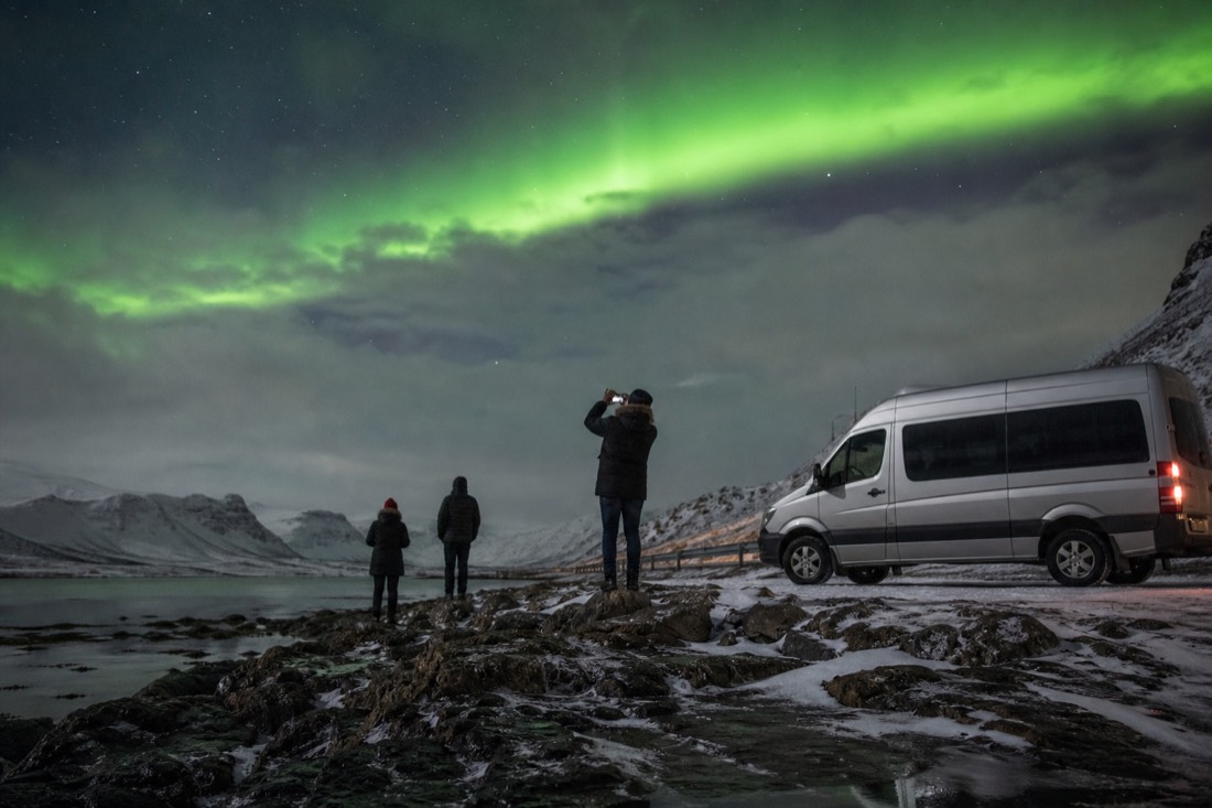 A guide photographing travelers under a green aurora band beside a minibus on a snowy roadside