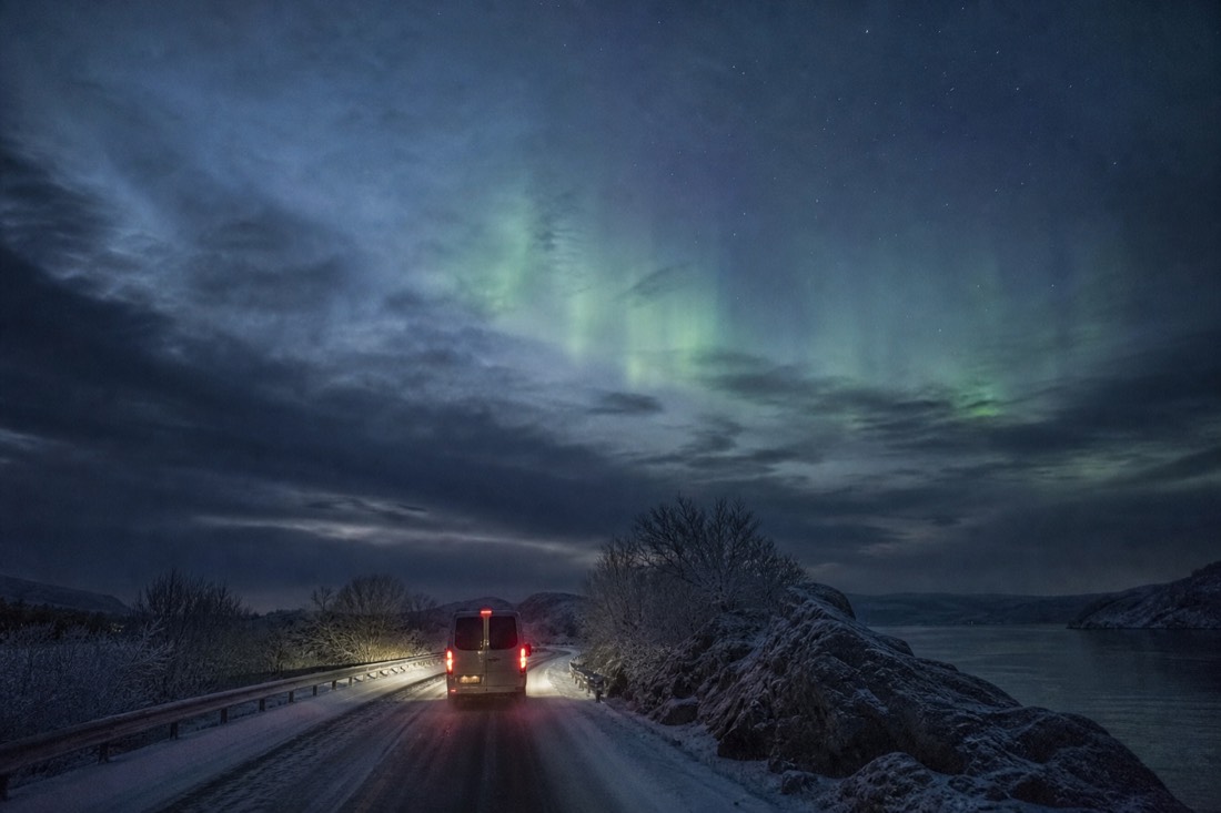 A minibus driving a snow-packed road at night with a faint aurora glow ahead near the Finnish border