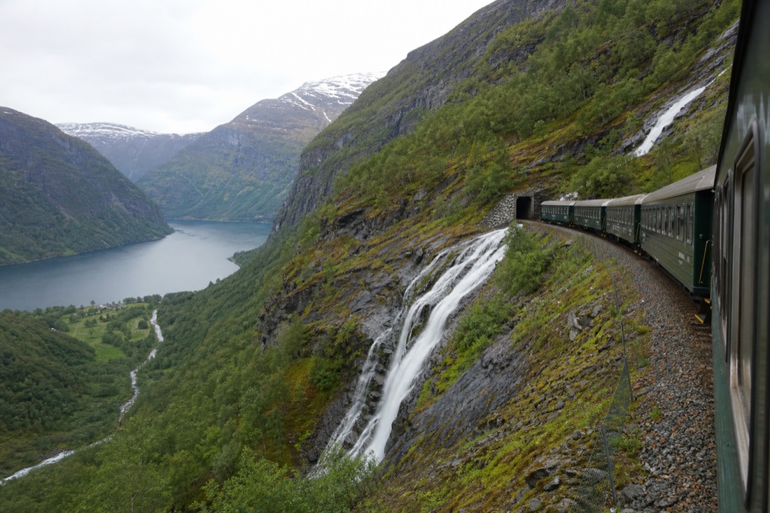 The Flam Railway's green carriages descending a steep valley wall past a waterfall toward the fjord
