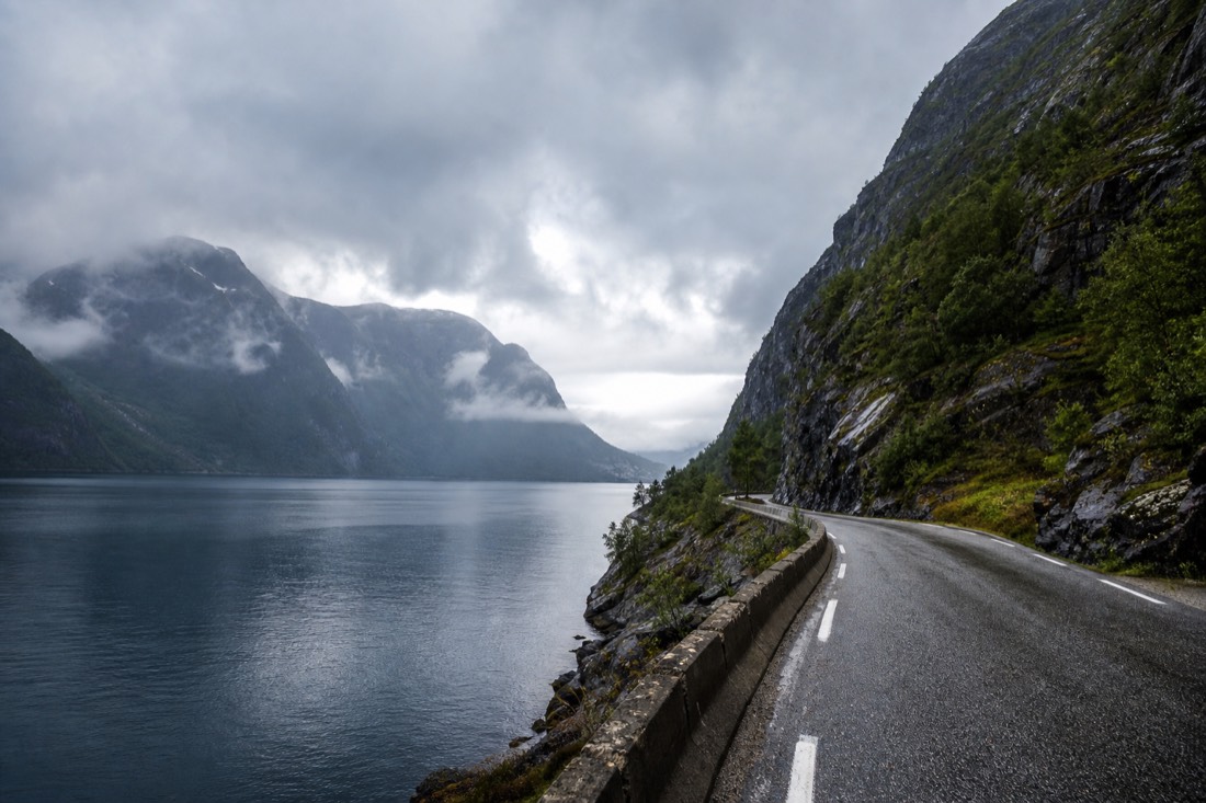 A coastal road winding between fjord water and steep mountains in western Norway under shifting light
