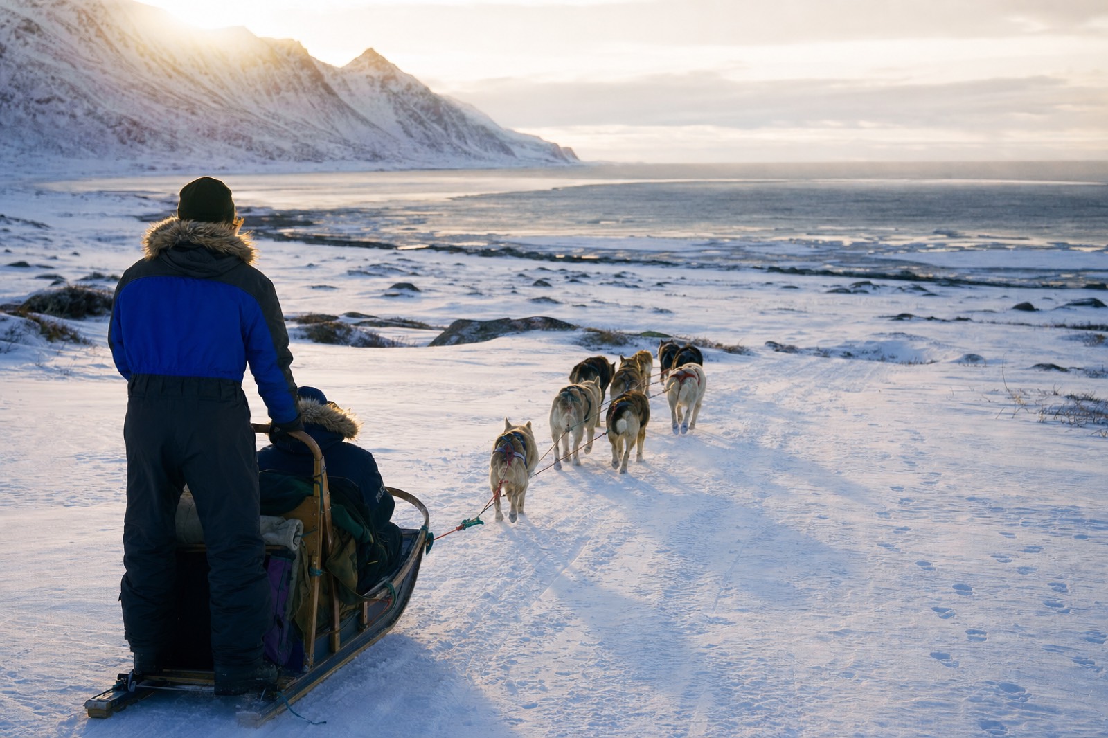 Two travelers driving a husky sled across an open snowfield with low winter sun on the mountains