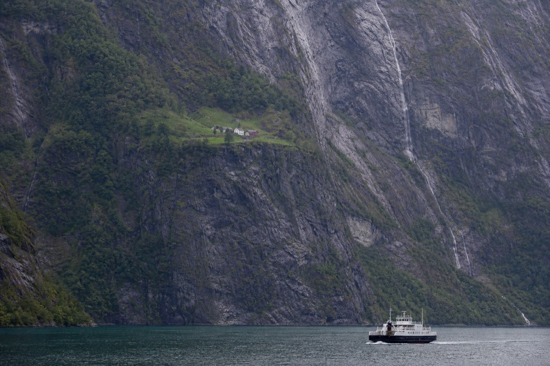 Steep-sided Norwegian fjord with a small ferry and a farm on a green ledge halfway up the cliff