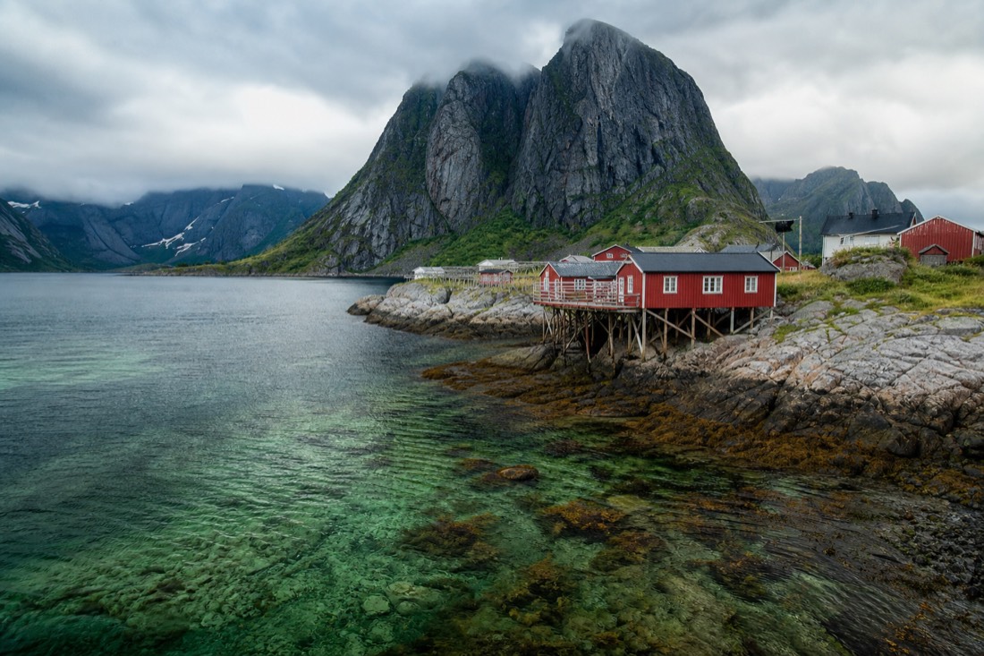 Red fishermen's cabins on stilts beside clear green water with sharp granite peaks rising directly behind