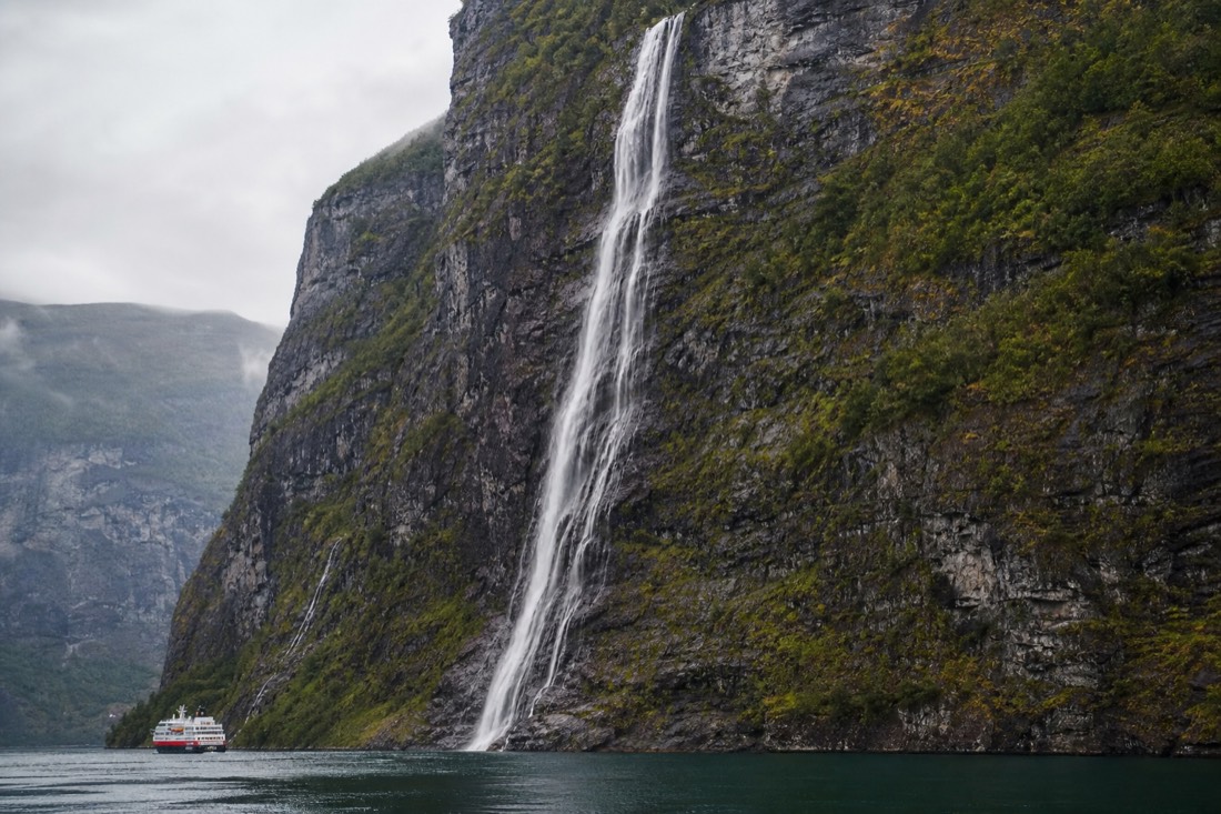 Tall thin waterfall spilling down a sheer green cliff into a narrow fjord with a small cruise boat below