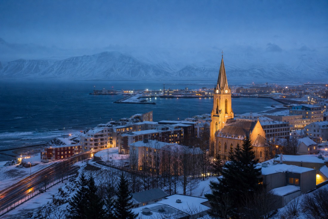 Snowy Arctic city street at blue hour with a lit cathedral spire and mountains across the dark water