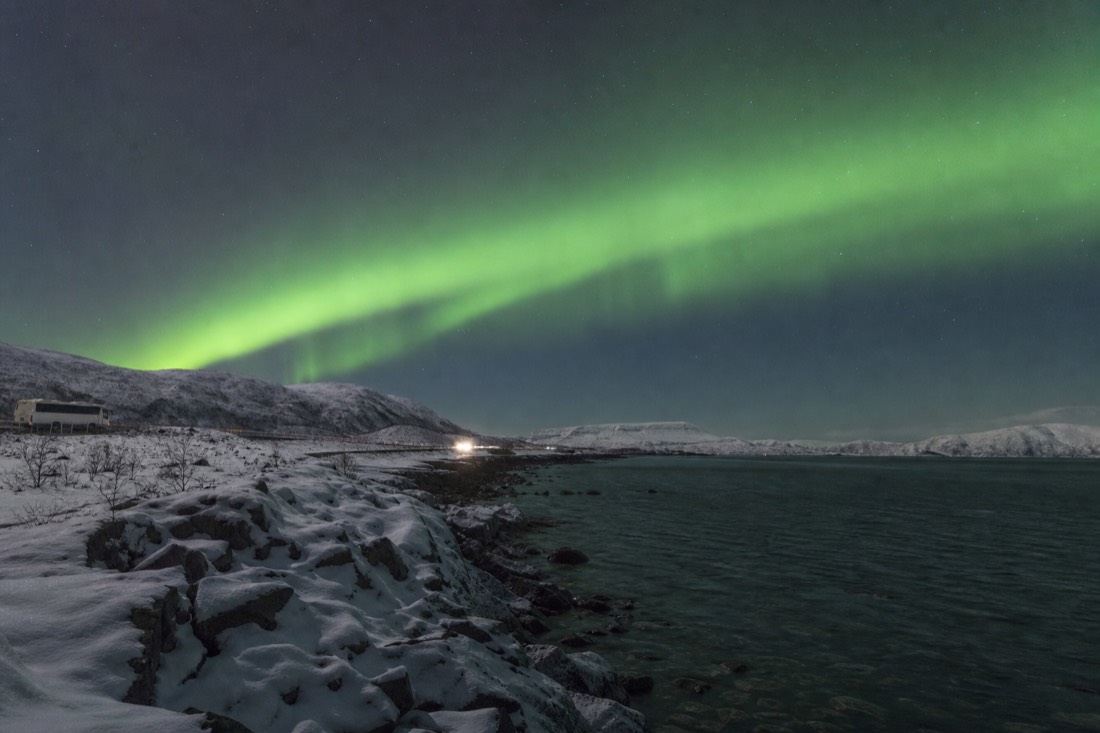 Green aurora arc over a snow-covered Arctic shoreline near Tromso with a parked minibus and headlamps in the distance