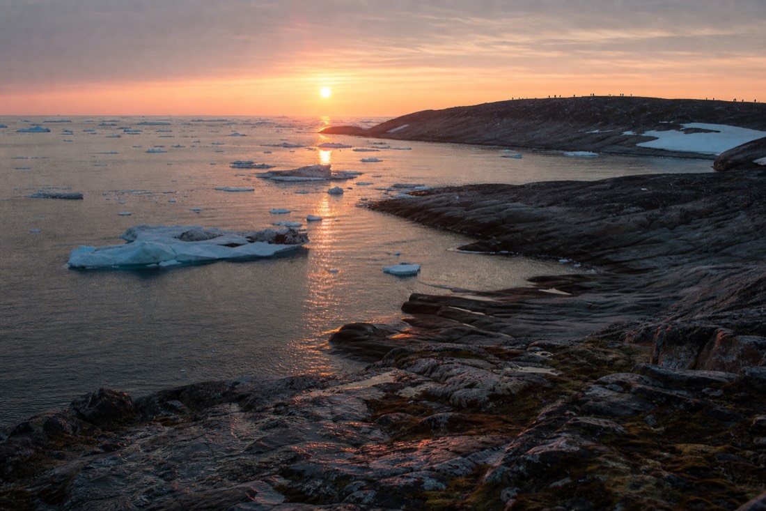 Low orange midnight sun over an Arctic coastline at one in the morning with hikers silhouetted on a ridge