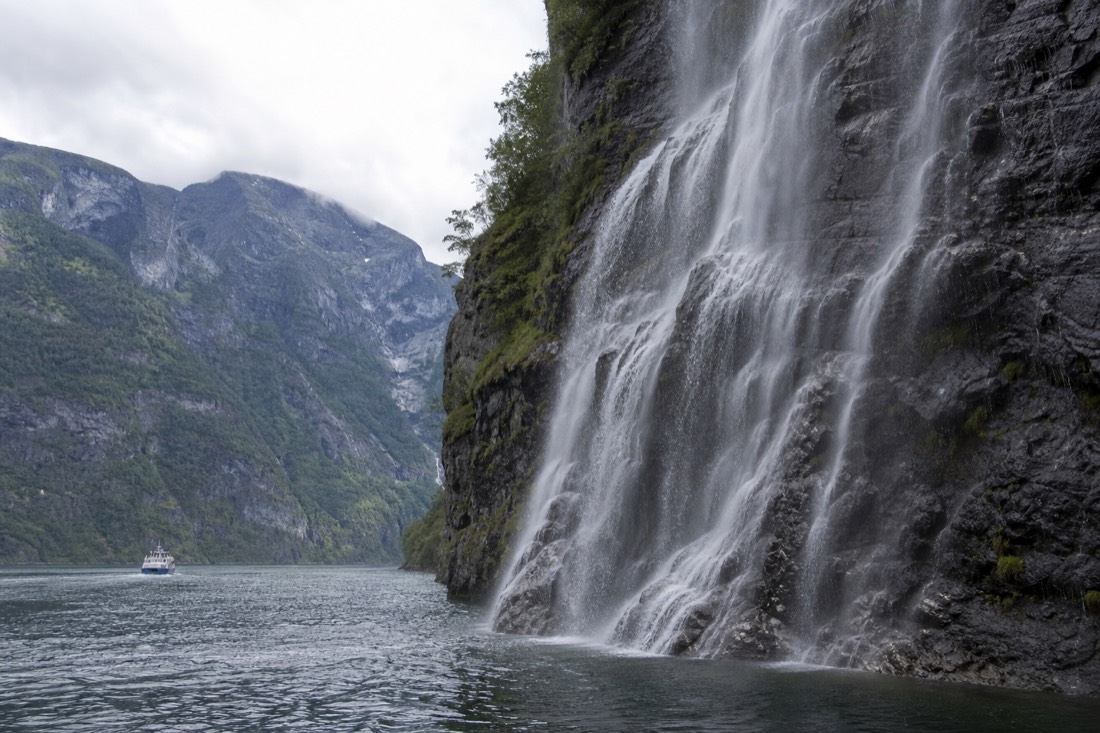 A small sightseeing boat passing close to a waterfall on a narrow fjord arm with steep green cliffs on both sides