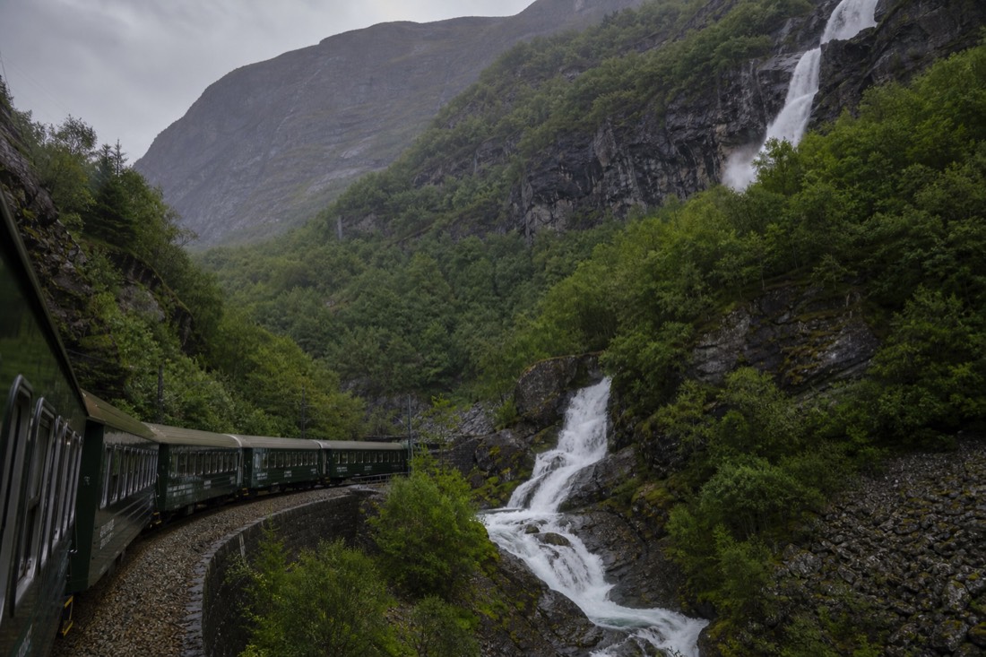 Green tourist train descending a steep valley past a waterfall on the Flam Railway line