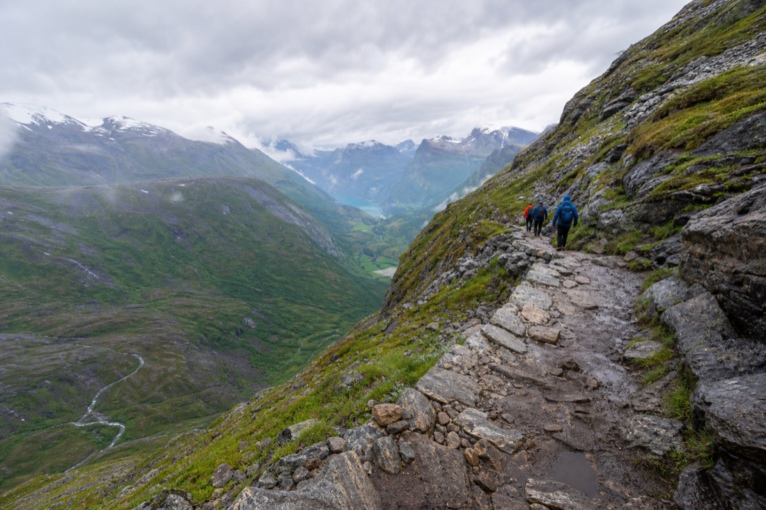 Hikers on a rocky mountain path above a green Norwegian valley with a turquoise lake far below