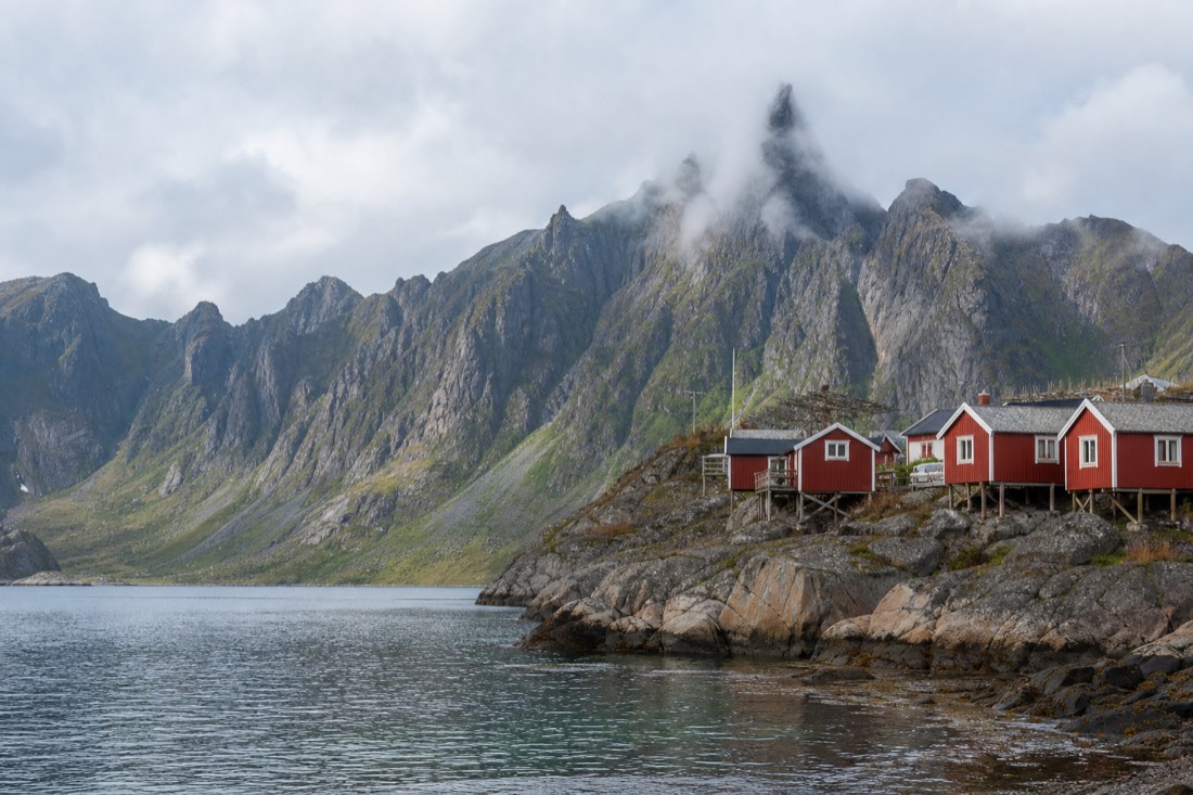 Red fishing cabins on stilts beside calm water with jagged Lofoten peaks behind