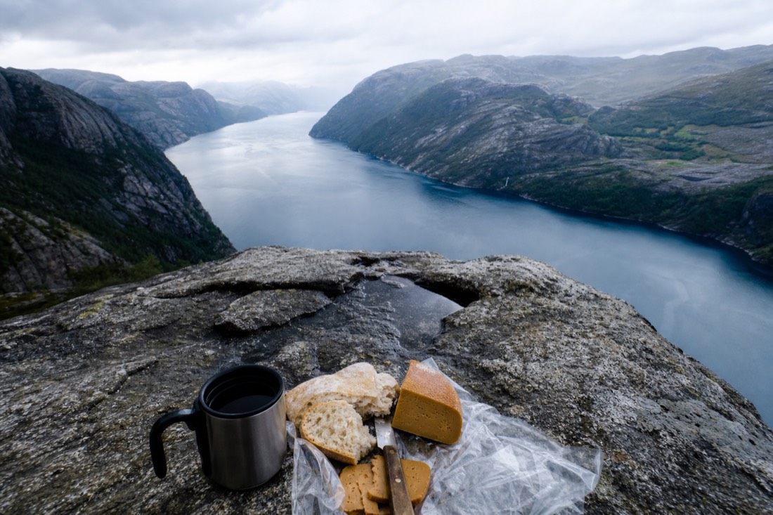 Supermarket picnic of bread, brown cheese and coffee laid out on a granite rock above a blue Norwegian fjord