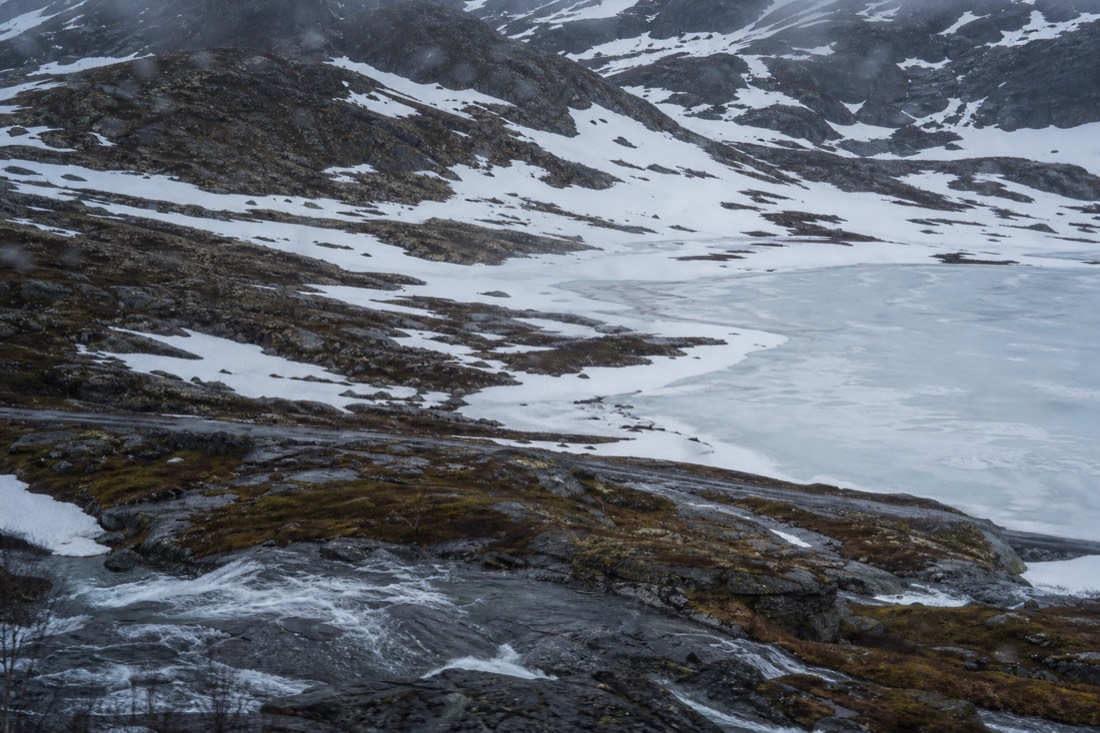 Train window view of snow patches and a frozen mountain lake on the Bergen Line above the treeline