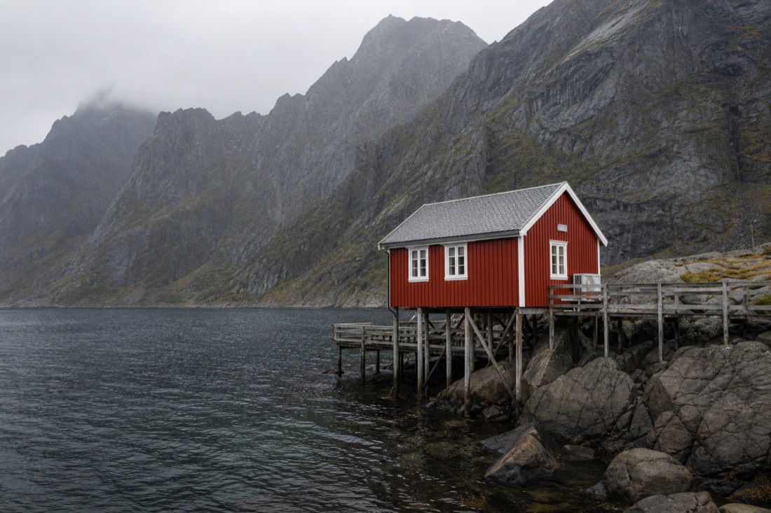 Small red-painted rorbu cabin on stilts above dark water with steep grey mountains behind