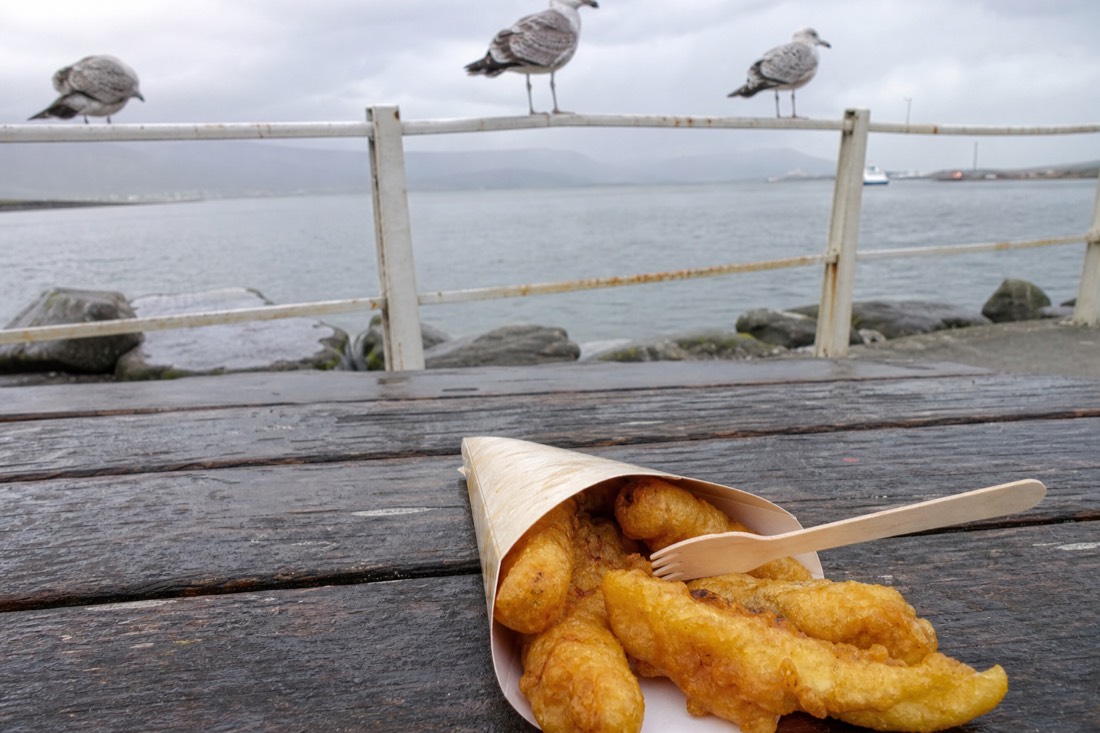 Paper cone of fried fish and a wooden fork on a harbourside bench with gulls on the railing