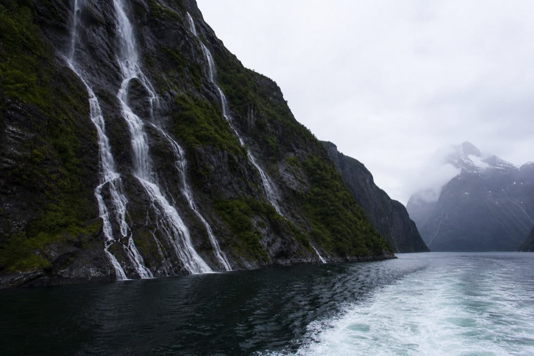 Small passenger boat cutting through a narrow fjord arm with waterfalls streaking the cliff face