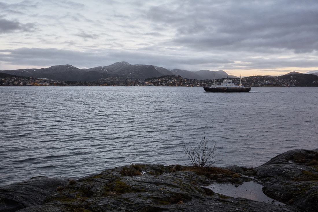 Coastal ferry crossing a wide grey fjord at dusk with the lights of a small town on the far shore