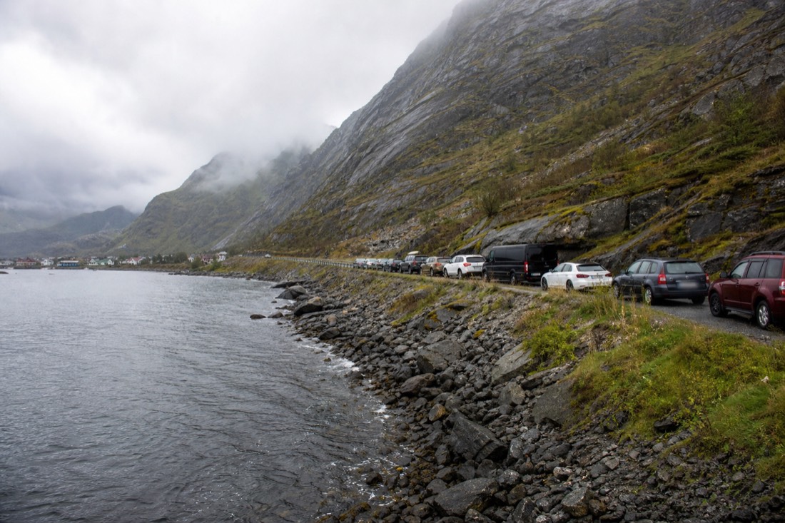 Parked cars lining a narrow roadside verge below a steep mountain in a Lofoten village
