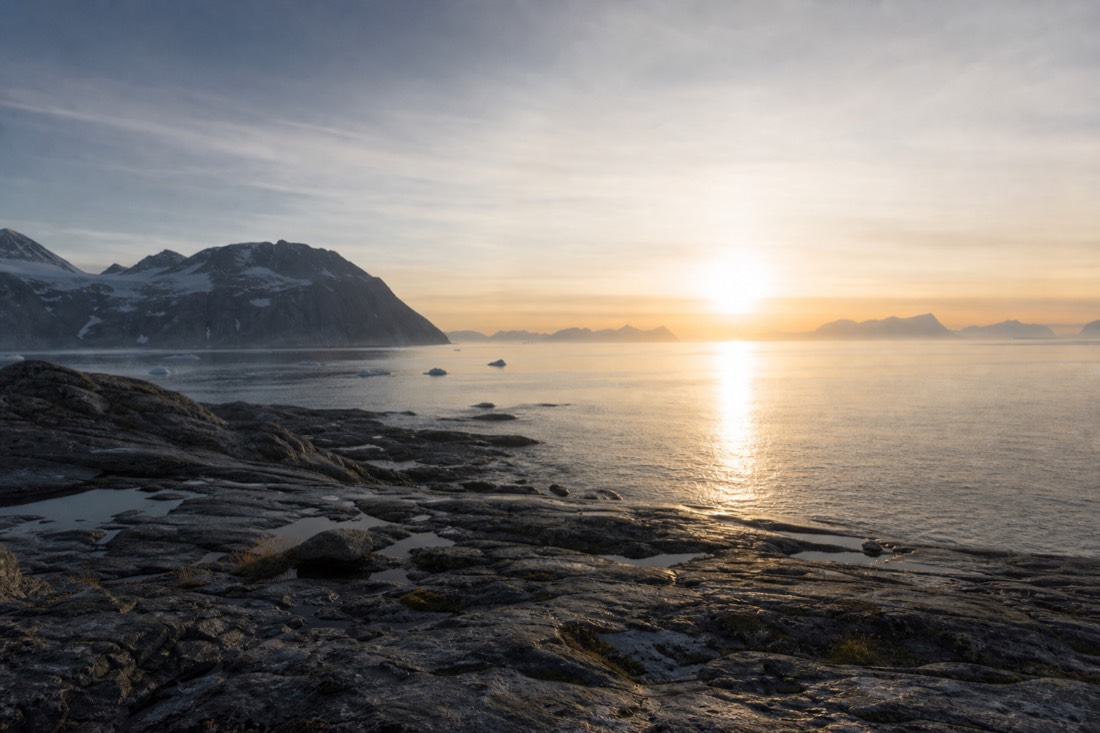 Sun hovering low over a still Arctic fjord at midnight with pale sky and dark rocky shoreline