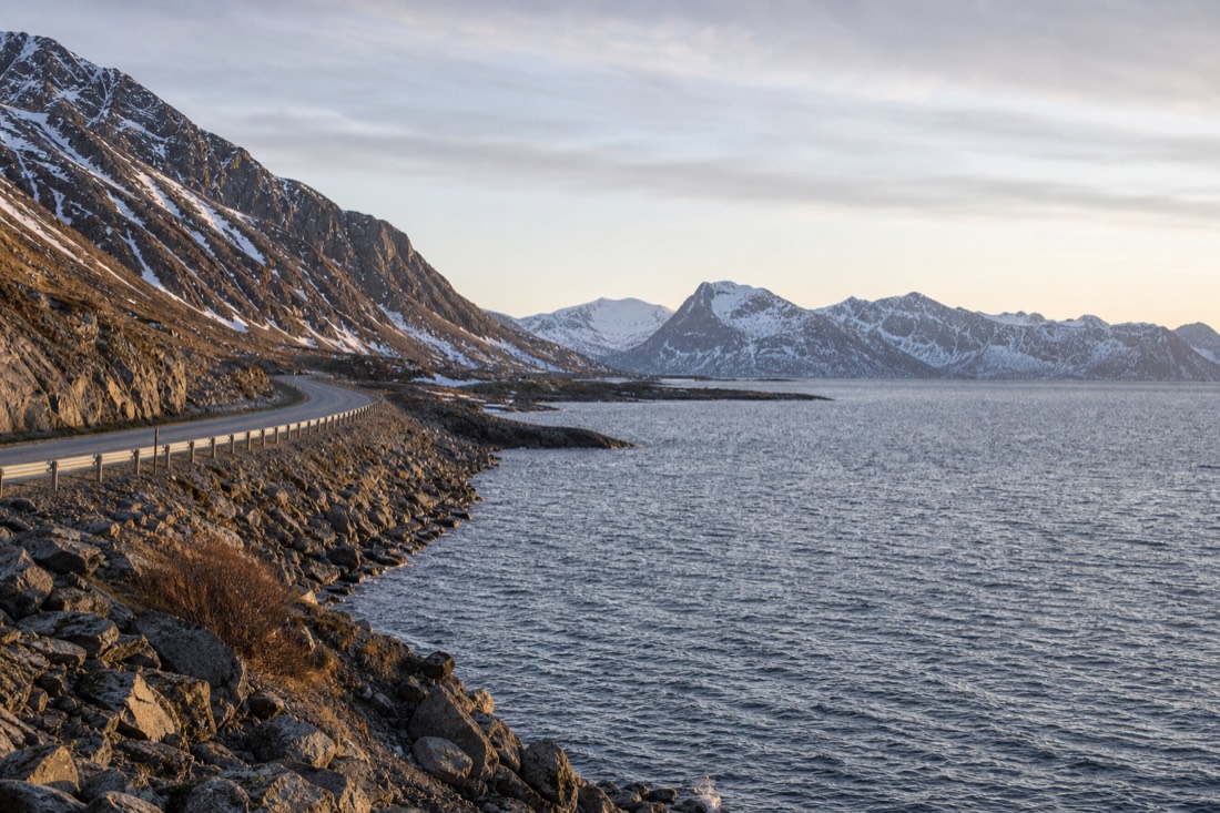 Coastal road in northern Norway lit by low midnight sunlight with snow patches on nearby peaks