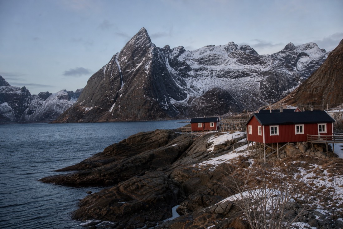 Red fishing cabins on a Lofoten shoreline under a pale night sky with jagged peaks behind