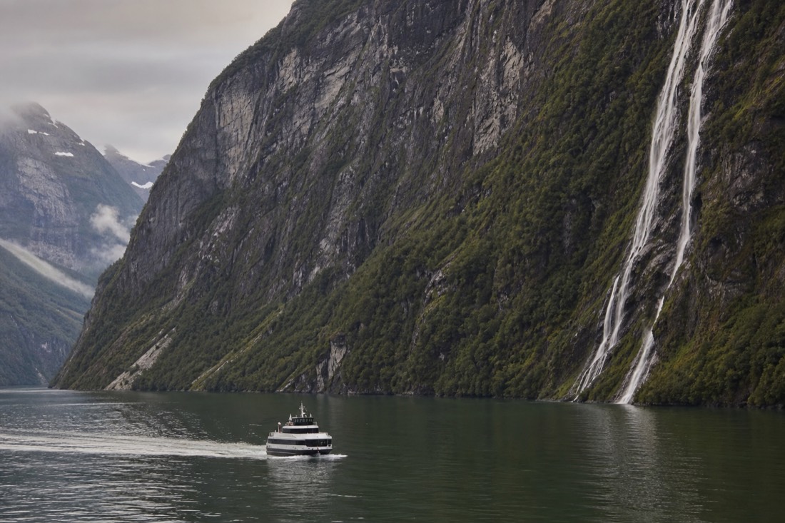 Sightseeing boat on a narrow green fjord in late evening light with waterfalls on the cliff walls