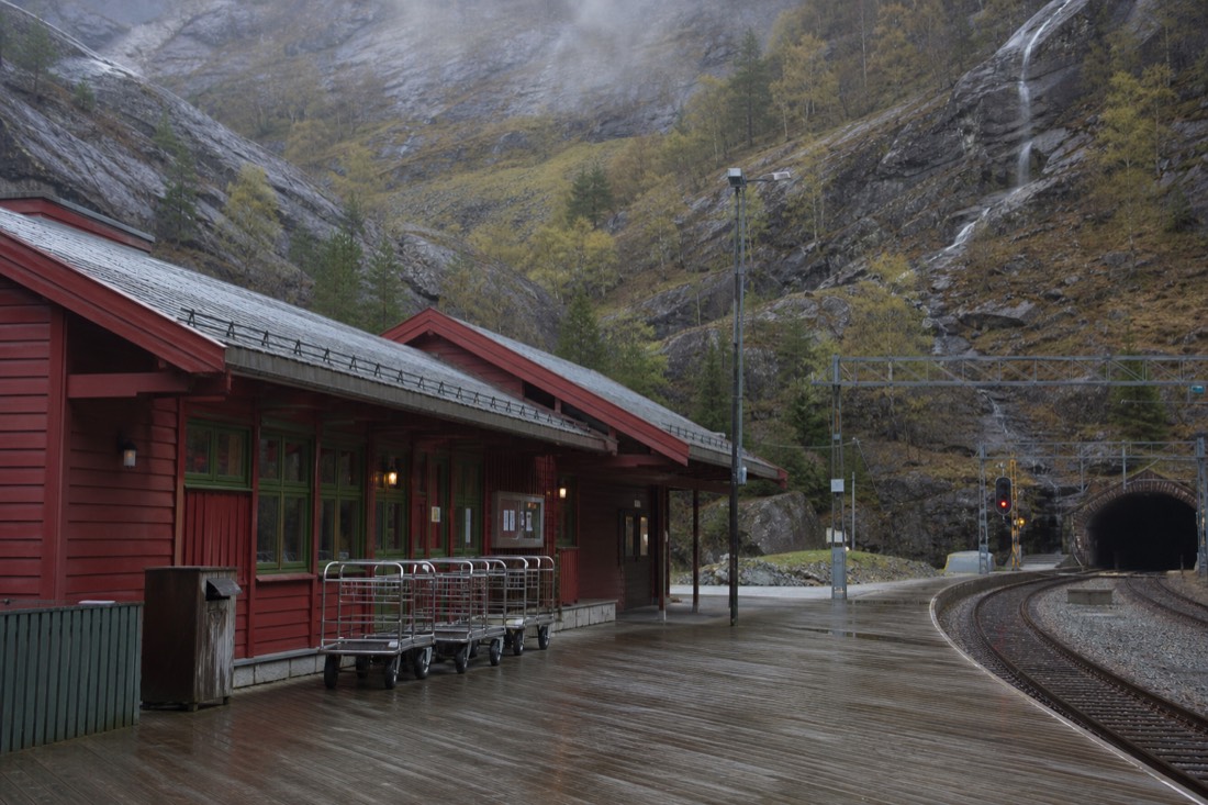 Wooden station platform at Flam with a red timber building, luggage trolleys and mountains rising behind