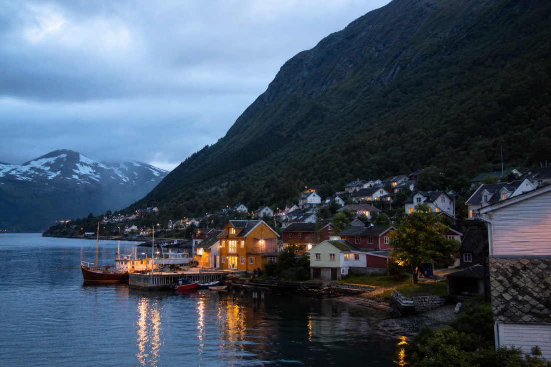 Small fjordside village at dusk with warm lit windows, a moored boat and steep dark mountains above