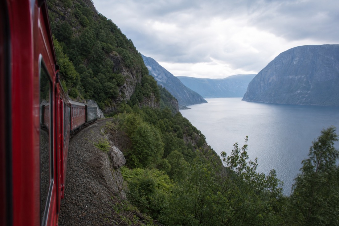 Red-painted railway carriage descending a steep green valley toward a fjord arm in western Norway